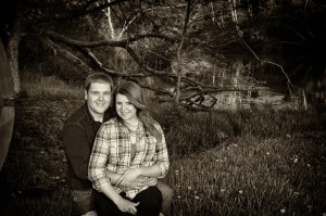 A young couple, Erika and Dan, smiling and embracing in a grassy area by a serene lake, surrounded by trees, in a sepia tone.