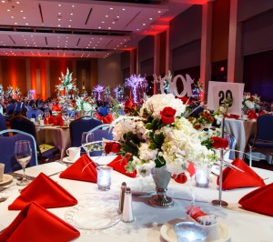 Elegant banquet hall set for the Centennial Event with tables adorned with red napkins, floral centerpieces, and glassware, under soft lighting.
