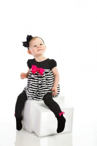 Toddler in a black and white polka dot dress with pink bows, engaging in a child portrait photography session, sitting on a white cube and looking up, against a white background.