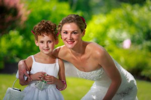 A bride and a young girl in white dresses smiling together in a garden at the Salem Cross Inn with lush greenery in the background.