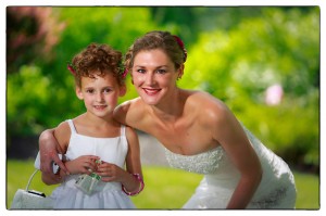 Laura and a young girl with curly hair, both smiling, pose in a garden during the Rich Wedding. Laura is wearing a white gown and the child holds a small purse.