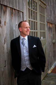 Man in a black suit and tie leaning against a wooden wall at the Salem Cross Inn, smiling and looking away.