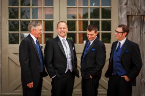 Four men in formal attire with blue accents smiling and standing together in front of a rustic wooden door with glass windows at the Salem Cross Inn.