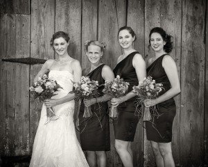 A black and white photo of a bride and three bridesmaids standing in front of a wooden door at Salem Cross Inn, holding bouquets, smiling at the camera.