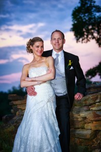 Laura and Rich posing together at sunset, standing next to a stone wall with a picturesque sky in the background.