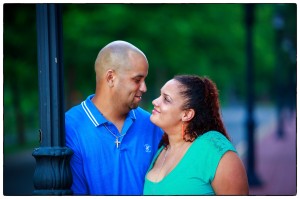 A couple lovingly gazing at each other, leaning on a lamp post in Forest Park, with a blurred green background. The man is bald and wearing a blue shirt, and the woman has curly
