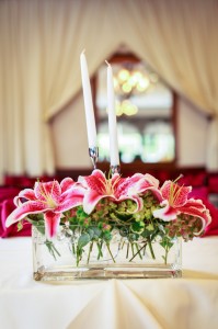Elegant table centerpiece featuring two white candles and a floral arrangement with wedding flowers including pink lilies in a glass vase, set in a festively decorated room.