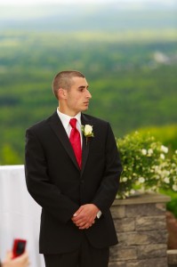 A man in a black suit and red tie stands thoughtfully at an outdoor wedding at The Log Cabin, with a scenic green valley in the background.