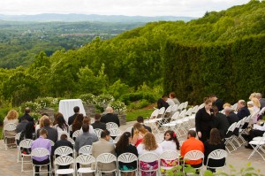 Outdoor wedding ceremony with guests seated on white chairs, overlooking a scenic hilly landscape, with a vine-covered wall in the background at The Log Cabin.