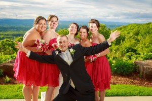 A groom kneeling in front and five bridesmaids in red dresses standing behind him, all smiling, with a scenic green hilly backdrop under a cloudy sky at The Log Cabin.