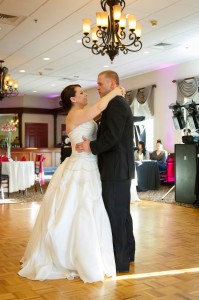 A bride and groom share a dance at The Log Cabin with guests and chandeliers in the background.
