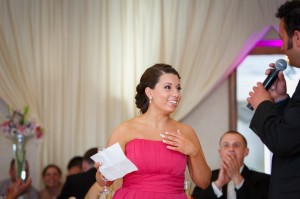 Alyssa in a pink dress smiling and touching her chest with a surprised expression at a Western Mass Wedding Photography event, as Jeff speaks into a microphone.