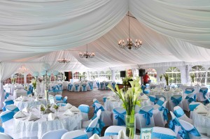 Elegant wedding reception setup under a large white tent with draped ceiling linens, round tables dressed in white and blue, and ornate chandeliers adorned with wedding flowers.