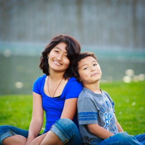 A young girl in a blue shirt and a young boy in a gray shirt sitting back to back on grass, smiling, during a child photo session, with a blurred green background.