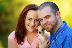 Brittany and Conrad holding a small chihuahua, smiling in an outdoor setting with brightly blurred greenery in the background during their engagement photos.