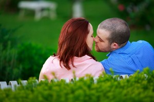 Brittany and Conrad kissing over a white fence surrounded by lush greenery during their engagement photo shoot.
