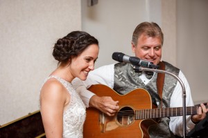 A smiling bride, Kara, in a beaded dress stands next to Brandon playing an acoustic guitar and singing into a microphone at their Easthampton Wedding Photography reception at The Log Cabin.