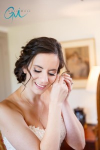 A bride in a strapless dress puts on an earring, smiling gently in a warmly lit room with a painting in the background at Salem Cross Inn.