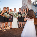 A bride in a white dress is taking a photo of six women in dark dresses holding bouquets on the sunny terrace of the Lord Jeffery Inn.