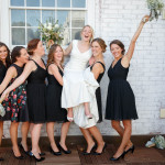A bride in a white dress laughing and lifting one leg, surrounded by six joyful bridesmaids in black dresses, holding bouquets against a brick wall at Lord Jeffery Inn weddings.
