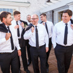 Group of men in formal attire laughing and adjusting their ties on a sunny outdoor terrace at Lord Jeffery Inn.