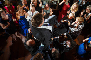 Groom being lifted by friends at a Salem Cross Inn wedding reception, surrounded by applauding guests in a dimly lit hall.