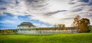 A panoramic view of an outdoor wedding venue with rows of white chairs facing a gazebo, captured by a wedding photographer, set against a backdrop of a cloudy sky and colorful autumn trees.