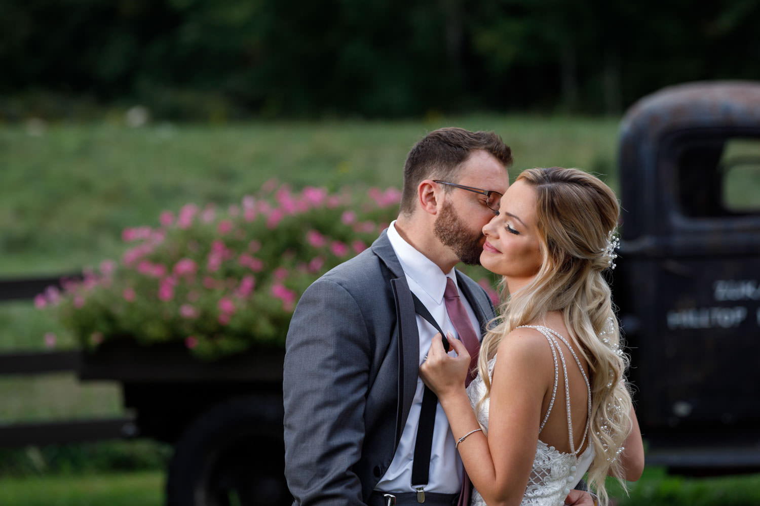 Groom kissing brides cheek while she pulls lightly on his suspenders with Zukas Hilltop Barn Truck in background