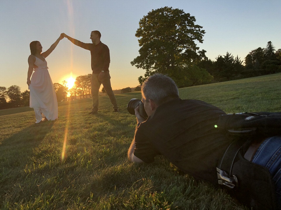 Photographer laying on the ground taking sunset photo of couple while guy twirls girl