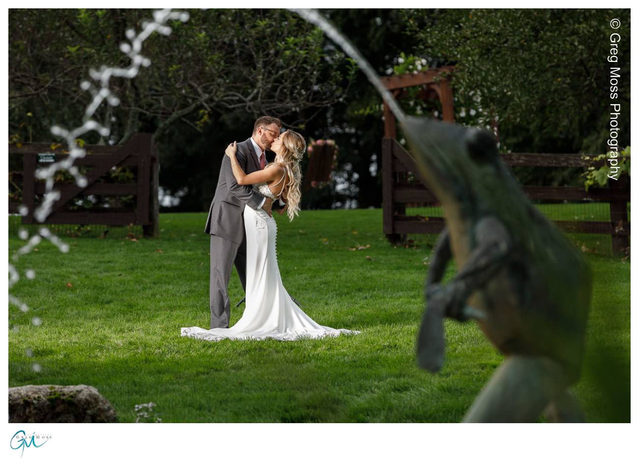 A bride and groom embracing and kissing in a lush garden at Zukas Hilltop Barn, with a statue and water spray in the foreground.