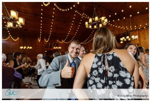 Man giving a thumbs-up at a lively wedding party at The Barn at Liberty Farms with string lights and guests dancing in the background.