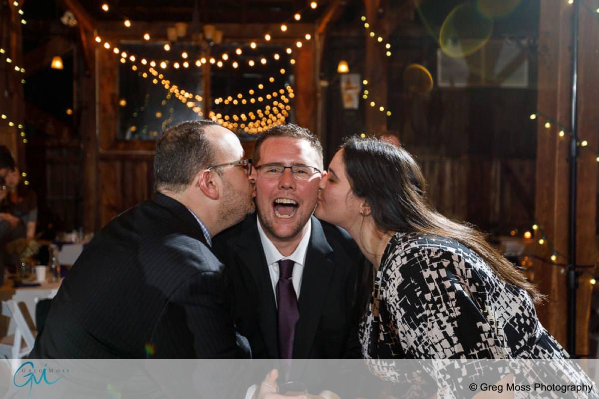 A man in glasses smiles joyfully as he receives kisses on each cheek from two people at a red barn wedding, with string lights in the background.