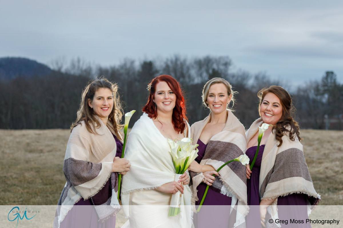 Four women in white dresses and draped shawls holding bouquets, smiling outdoors with a field and mountains in the background at Red Barn Weddings.