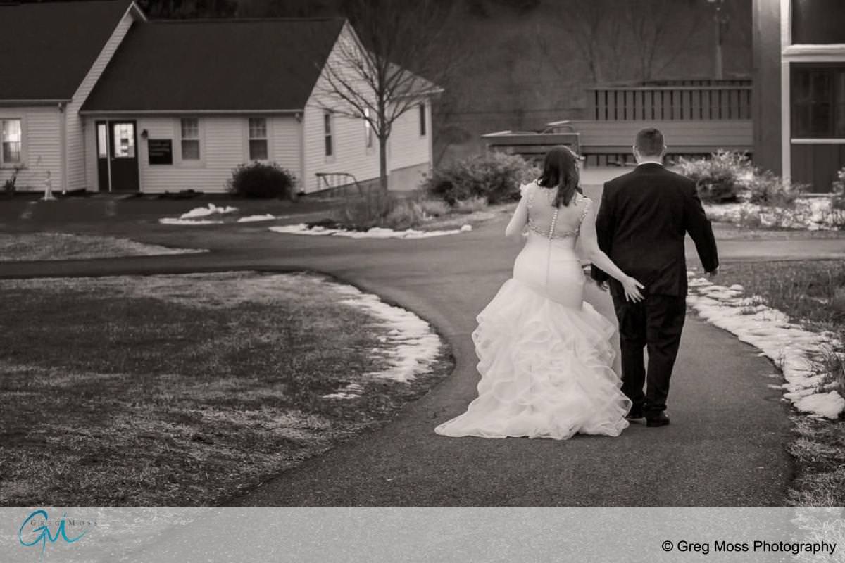 A bride in a white gown and a groom in a black suit walk hand in hand on a curved pathway, with houses, a red barn, and patches of snow in the background. Black and white