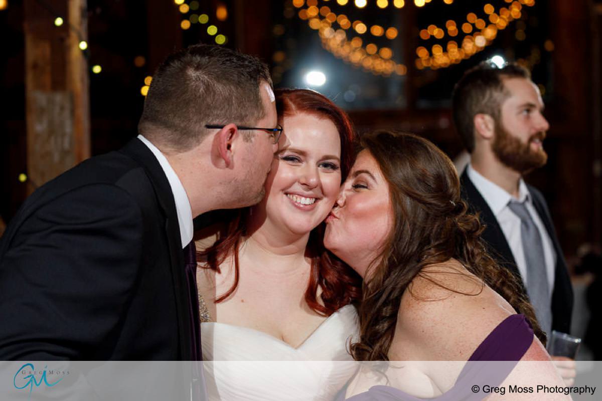 A bride smiles as she receives kisses on the cheek from a man and a woman at a Red Barn Wedding, with another man looking on in the background.