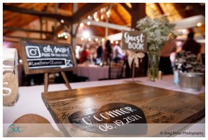 A wooden guest book with signatures at a Barn at Wight Farm wedding reception, with a chalkboard sign in the foreground, in a rustic venue with guests in the background.