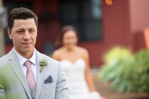 Groom in a gray suit standing in focus, with the bride in a white dress slightly blurred in the background, standing against a red barn at Wight Farm.