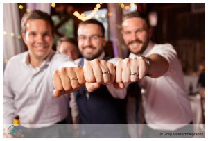 Three smiling men at a Hampshire College wedding, showing their fists with matching rings to the camera, with soft-focused lights in the background.