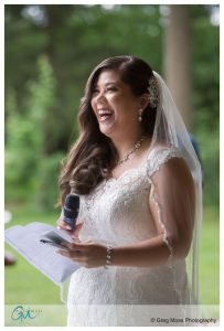 A joyful bride in a lace dress and veil holding a microphone and reading from a paper at Look Park, with trees in the background.