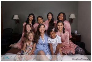 A group of nine diverse women and girls, dressed in robes and casual outfits, smiling and posing together in a hotel room for a Look Park wedding.