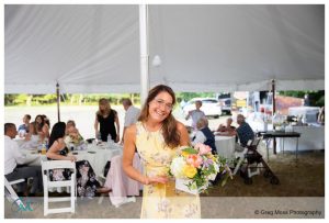 A woman in a yellow dress smiling while holding a bouquet at an outdoor farm wedding reception.