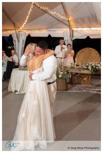 A newlywed couple shares an emotional dance under a tent with string lights at a Plimouth Museum Wedding, surrounded by seated guests and a floral-decorated table in the background.
