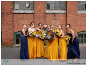 A joyful Boylston Rooms wedding group posing outdoors, where a bride in white is playfully lifted by three bridesmaids dressed in yellow, alongside three others in navy blue, all holding sunflower bou