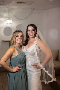 Two women smiling at a log cabin wedding, one in a green bridesmaid dress and the other in a white lace bridal gown.