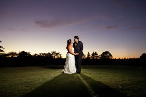 A couple in wedding attire embraces on a grassy field at Crestview CC during sunset, with a vibrant sky and long shadows stretching behind them.