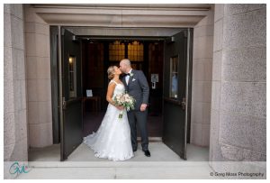 A bride and groom kiss in a doorway, framed by stone walls, with the groom holding a bouquet during their Holyoke wedding.
