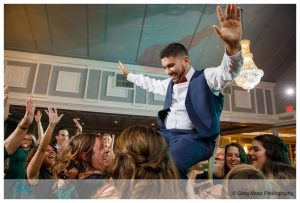 A man in a blue vest and white shirt crowd surfing over a group of excited guests at a lively wedding at Glastonbury Country Club.