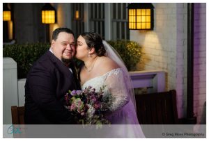 A bride and groom embracing and kissing outdoors at the Inn on Boltwood, with the bride holding a bouquet of flowers.