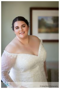 A bride in an off-the-shoulder lace wedding dress smiles gently, standing in a room with a landscape painting in the background at the Inn on Boltwood fall wedding.
