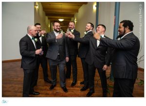 A group of men in suits, holding drinks, cheerfully toasting at a Mill 1 wedding in Holyoke, MA.